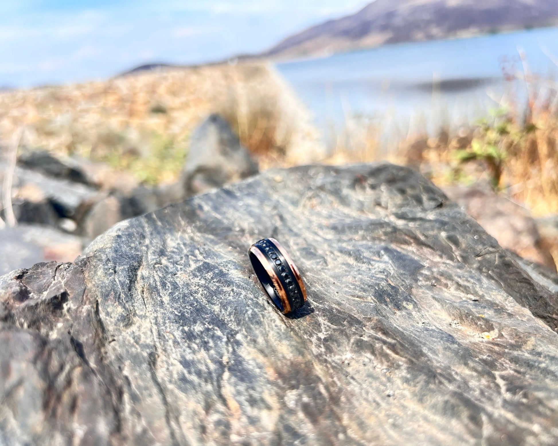 Black & Rose gold ring with CZ's on a rock with a scenic background of water and mountains