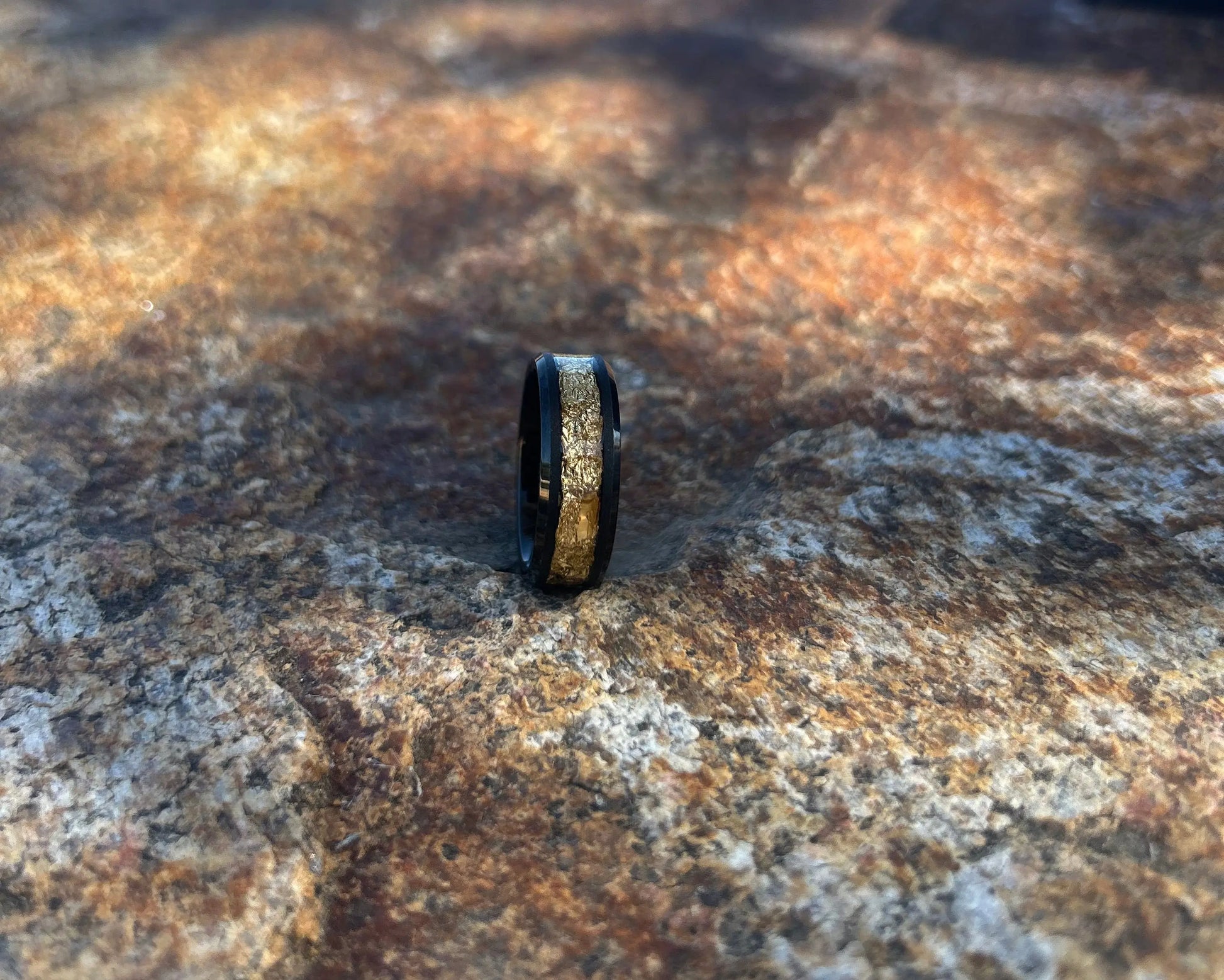 Black ring with gold inlay on a textured stone surface with a blurred blue sky background